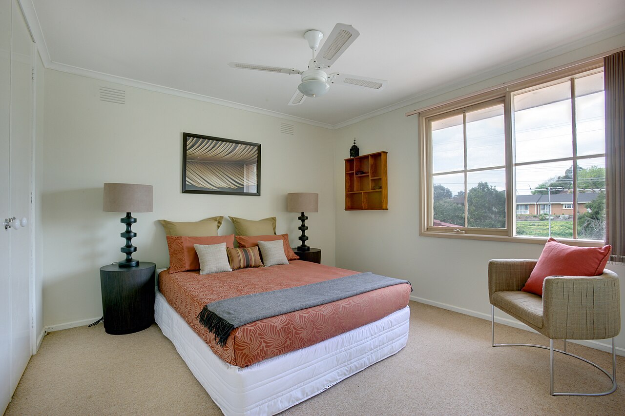 Bright lived-in bedroom with a ceiling fan and wall air vents above the bed