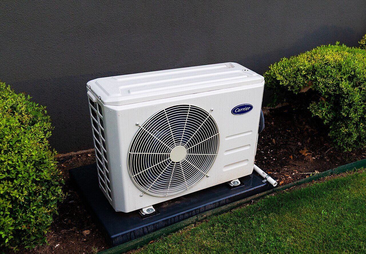 Wall-split heat pump outdoor unit set in a garden bed against dark cladding