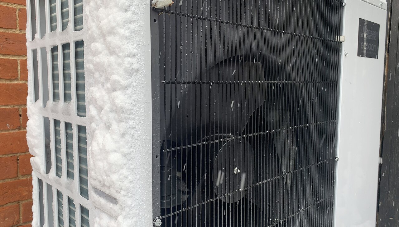 Heat pump outdoor unit running in falling snow against a brick wall